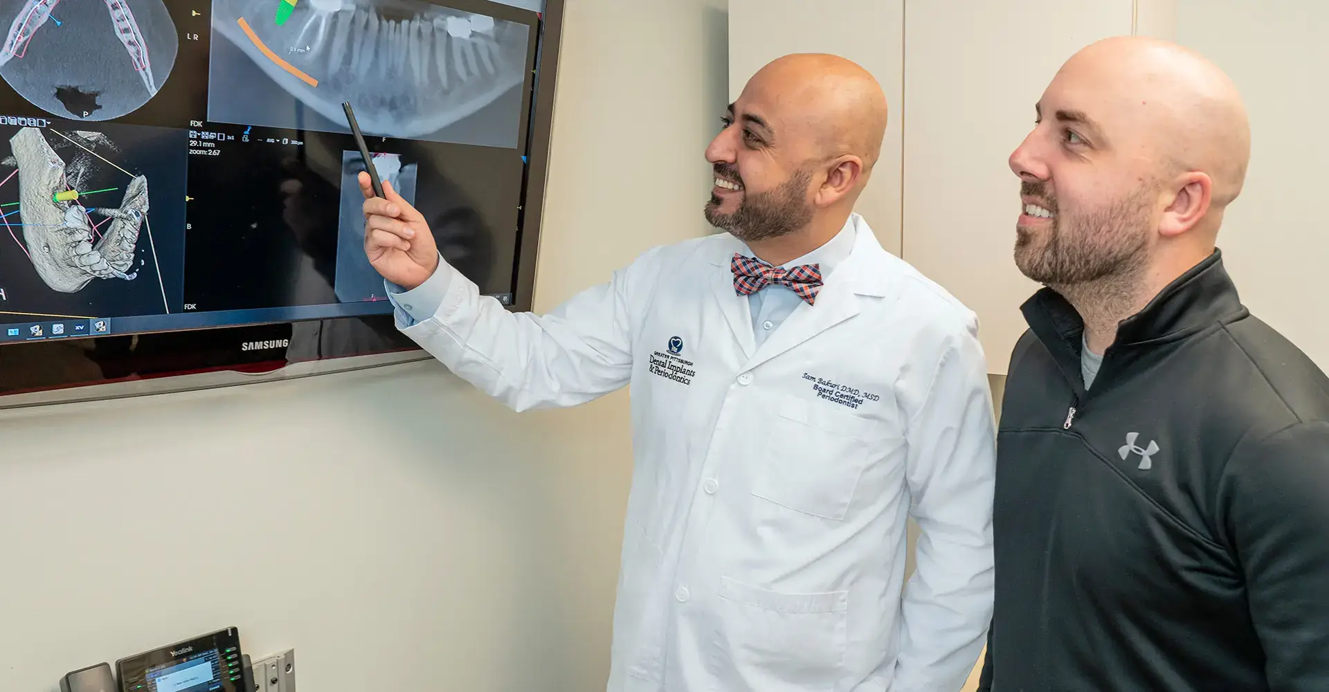 A dentist in a white coat, smiling, points at dental X-rays on a screen while explaining something to a patient. Both are standing in an examination room. The patient, wearing a black jacket, also smiles while listening.