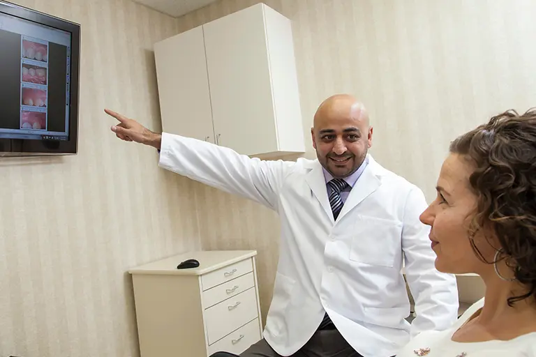 A doctor in a white coat is happily explaining medical images displayed on a wall-mounted monitor to a seated patient in an exam room. The patient listens attentively.